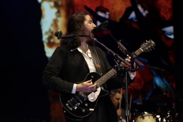 A man sings with a black guitar on a stage with projections behind him.